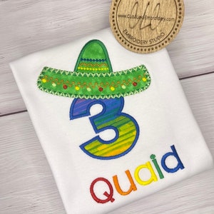 May include: A white t-shirt with a rainbow embroidered number 3, a sombrero, and the name "Quaid". The shirt is on a wooden surface. The text "Cuba Lake Crafts" and "www.CubaLakeEmbroidery.com" is on a wooden circle.