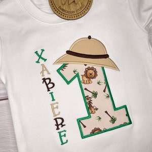 May include: White embroidered baby onesie with a green number one, a safari hat, and the name "XAVIER" embroidered vertically on the left side. The number one is filled with a jungle print featuring lions and palm trees.