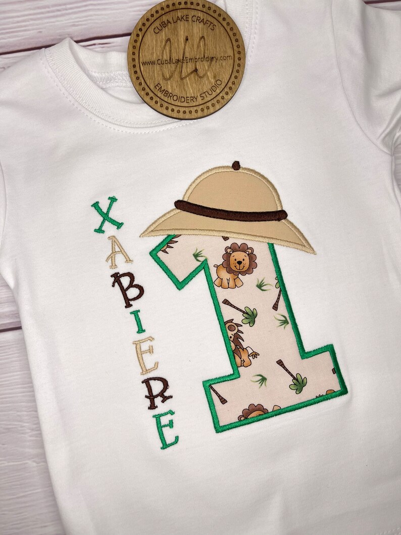 May include: White embroidered baby onesie with a green number one, a safari hat, and the name "XAVIER" embroidered vertically on the left side. The number one is filled with a jungle print featuring lions and palm trees.
