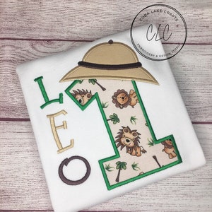 May include: White t-shirt with a green and brown embroidered number one. The number one is surrounded by a jungle theme with palm trees and lions. The letters "LEO" are embroidered in brown on the left side of the shirt. A brown safari hat is embroidered above the number one.