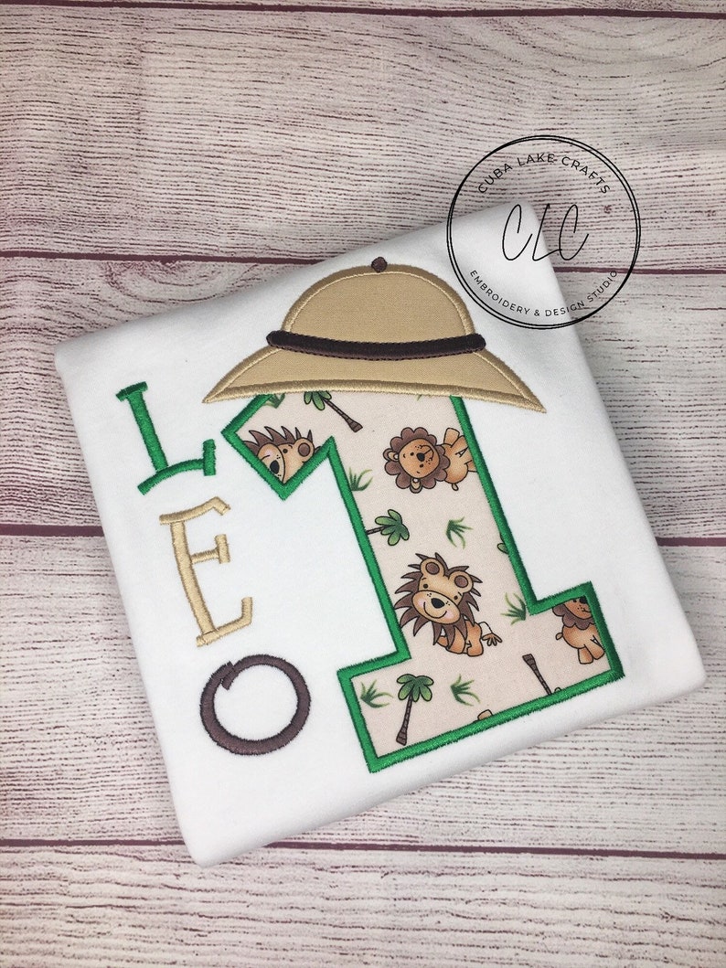 May include: White t-shirt with a green and brown embroidered number one. The number one is surrounded by a jungle theme with palm trees and lions. The letters "LEO" are embroidered in brown on the left side of the shirt. A brown safari hat is embroidered above the number one.