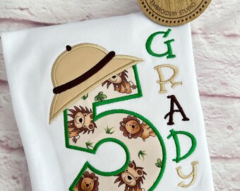 Può includere: Maglietta bianca con un numero 5 verde, un cappello da safari marrone e il nome "Grady" ricamato in marrone e giallo. Il numero 5 è riempito con un motivo di leoni marroni e foglie verdi.