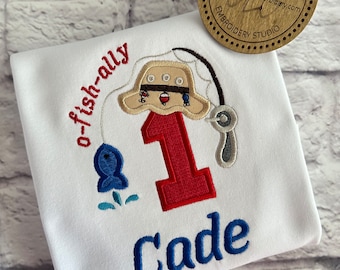 May include: White onesie with embroidered fishing themed design. The design includes a red number one, a blue fish, a fishing rod, and a hat. The name "Cade" is embroidered below the number one.