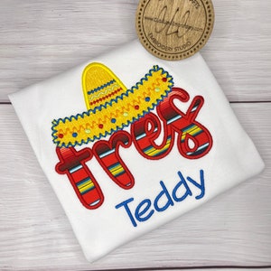 May include: White t-shirt with embroidered red, yellow, and blue "tres" with a yellow sombrero on top. The name "Teddy" is embroidered in blue below the "tres".