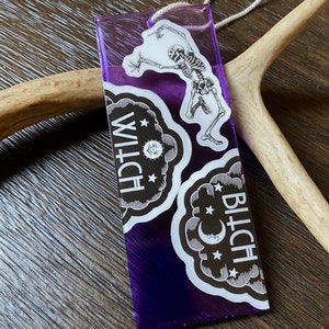 May include: Purple plastic tag with a skeleton dancing and two white stickers with the words "WITCH" and "BITCH" in black text.