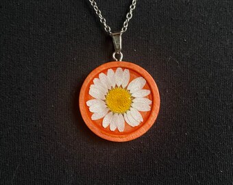 May include: A silver chain necklace with a round pendant featuring a white daisy pressed into a red wooden circle.