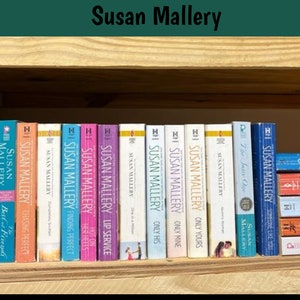 May include: A collection of Susan Mallery books displayed on a wooden shelf. The spines of the books are visible, showcasing titles like "Yours Truly," "Summer Days," and "Almost Perfect." The books are in various colors, including orange, pink, and blue.
