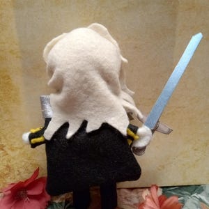 May include: A handmade felt doll of a figure in a black outfit with yellow and black accents, holding a silver and blue sword. The doll has white hair and a white collar. The background is a floral pattern and a tan wall.