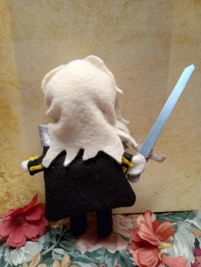 May include: A handmade felt doll of a figure in a black outfit with yellow and black accents, holding a silver and blue sword. The doll has white hair and a white collar. The background is a floral pattern and a tan wall.