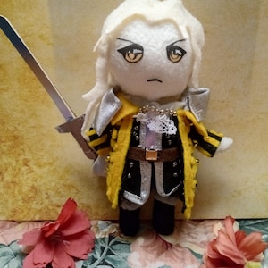 Puede incluir: Muñeco de peluche hecho a mano de un personaje con cabello blanco, que lleva un abrigo amarillo y negro con detalles plateados y sostiene una espada plateada. El muñeco tiene una expresión seria y está decorado con pequeñas cuentas. El fondo presenta un estampado floral.