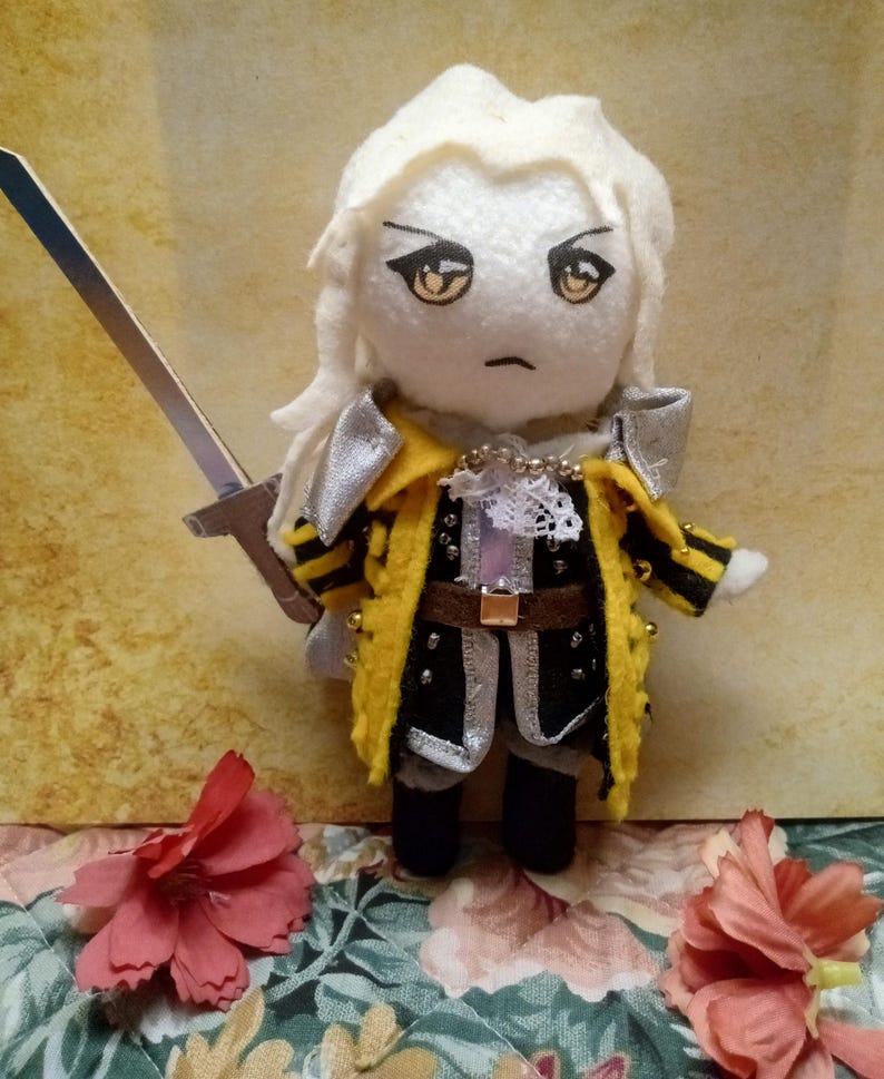 May include: A handmade plush doll of a character with white hair, wearing a yellow and black coat with silver accents, and holding a silver sword. The doll has a stern expression and is decorated with small beads. The background features a floral pattern.