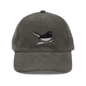 May include: A corduroy baseball cap in a muted green color. The cap features an embroidered design of a black and white bird perched on a branch. The bird has a yellow eye and is facing right.