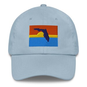 May include: Light blue baseball cap with an embroidered design of the Florida state outline over a colorful background. The background features horizontal stripes of orange, yellow, and blue. The cap has a curved brim and a button on top.