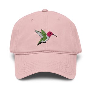 Op de afbeelding: Een lichtroze baseballpet met een geborduurd ontwerp van een vliegende kolibrie. De vogel heeft een felroze kop, een groen lichaam en witte en grijze vleugels. De pet heeft een gestructureerde kroon, een gebogen klep en een knoop bovenop.