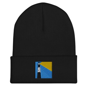 Puede incluir: Gorro de punto negro con un pequeño parche bordado de un faro sobre un fondo amarillo y azul.