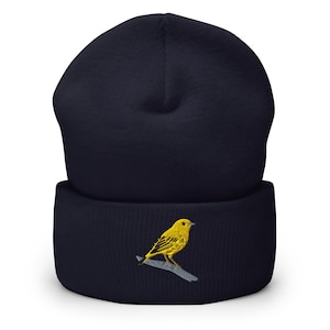 May include: A dark navy blue cuffed beanie featuring an embroidered design of a bright yellow bird perched on a light grey branch. The knit hat has a solid colour construction with a textured surface and a prominent fold-over cuff at the bottom.