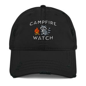Pode incluir: Boné de beisebol preto desgastado com as palavras "CAMPFIRE WATCH" bordadas em branco. Um guaxinim e um gráfico de fogueira estão centralizados acima do texto. O boné tem uma aba curva e um visual desgastado.