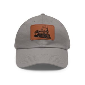 May include: Gray baseball cap with a brown leather patch featuring a black illustration of a vintage steam train. The cap has a curved brim and a classic six-panel design, suitable for casual wear.