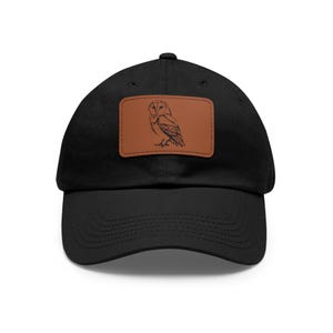 Op de afbeelding: Zwarte baseball cap met een bruin leren patch met een gedetailleerde schets van een uil. De uil is in profiel afgebeeld en toont zijn kenmerken. De cap is gemaakt van een zacht materiaal en heeft een gebogen klep.