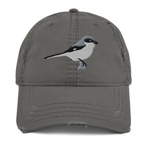 May include: A distressed gray baseball cap featuring an embroidered bird design. The bird is depicted in shades of gray, black, and white. The cap has a curved brim and a distressed, worn appearance, with visible fraying around the edges.