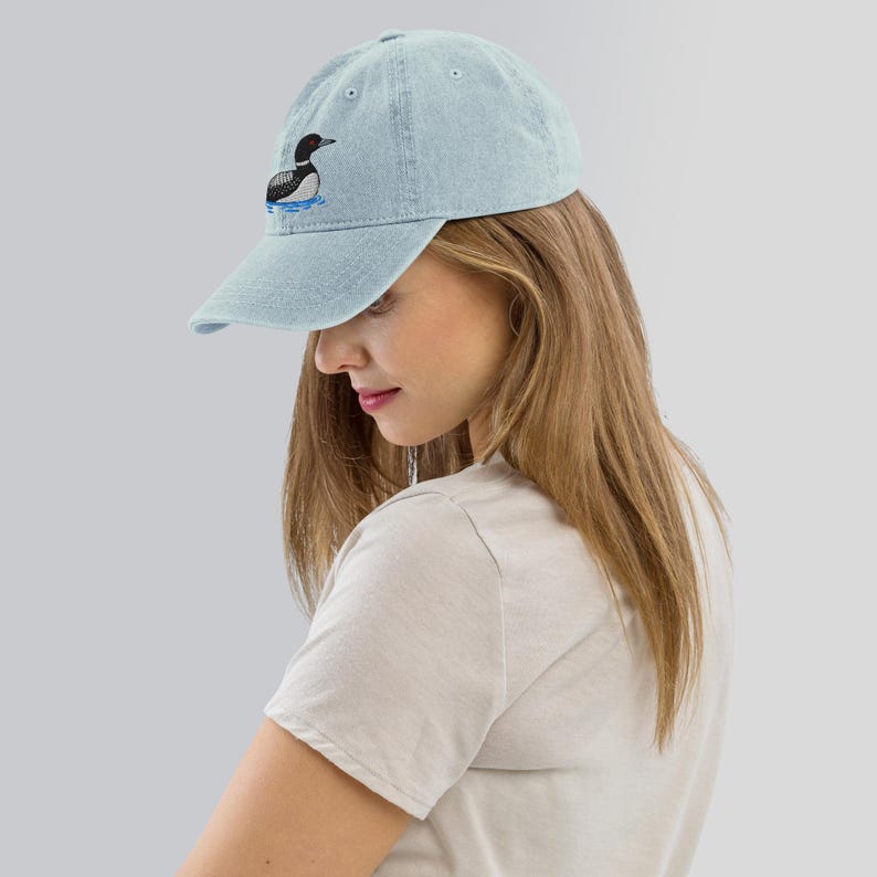 May include: A light blue denim baseball cap with an embroidered black and white loon design. The loon is facing left and is swimming in blue water.