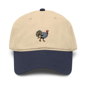 Könnte beinhalten: Eine beige Baseballkappe mit dunkelblauem Schirm und einer kleinen gestickten Grafik eines Truthahns auf der Vorderseite. Der Truthahn hat graue Federn, einen roten Kehllappen und braune Schwanzfedern und ist mittig auf der Kappe platziert.