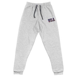 Puede incluir: Pantalones deportivos grises claros con cintura con cordón y la palabra "USA" bordada en la pierna izquierda en rojo, blanco y azul. Los pantalones deportivos están hechos de un material suave y cómodo.