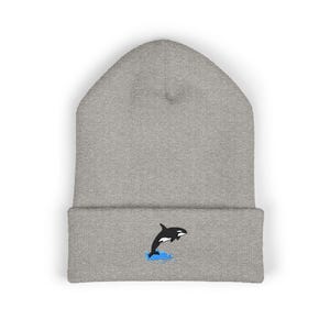 Op de afbeelding: Een grijze beanie met een geborduurd ontwerp van een zwart-witte orka die uit blauw water springt. De beanie heeft een omgeslagen manchet en een zachte, gebreide textuur. Het orka-ontwerp is gecentreerd op de manchet.