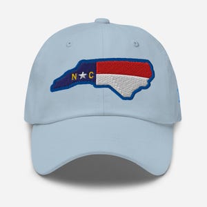 May include: A light blue baseball cap featuring an embroidered patch of the North Carolina state outline. The patch displays the state flag design with a blue section containing a white star and the yellow letters N and C, plus red and white stripes.