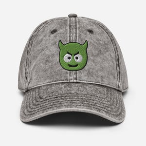 May include: A gray, vintage-style baseball cap with a curved brim. The cap features an embroidered green cartoon devil face with white eyes and a smiling mouth. The cap is made of a textured fabric.