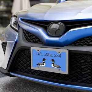 Può includere: Targa automobilistica con sfondo blu e la scritta "LIVE. LAUGH. LOON". Due illustrazioni di uccelli colymbus sono sotto il testo. La targa è montata sulla parte anteriore di un'auto blu.