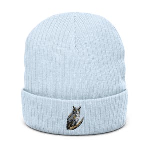 May include: A light blue ribbed knit beanie featuring a small, detailed embroidered graphic of a grey and brown owl perched on a branch, centered on the folded cuff of the hat. The beanie has a vertical ribbed texture throughout the fabric.
