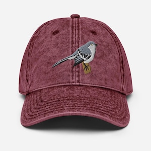 Op de afbeelding: Een bordeauxrode vintage gewassen denim baseballpet met een geborduurde grijze en witte vogel op de voorkant.