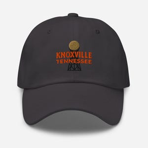 Puede incluir: Gorra de béisbol gris carbón con el texto bordado "KNOXVILLE TENNESSEE" en naranja. El diseño incluye un elemento redondo y texturizado encima del texto y una estructura estilizada debajo.