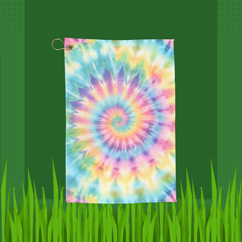 May include: A tie-dye golf towel with a vibrant spiral pattern in shades of blue, purple, yellow, and pink. The towel has a metal grommet and a loop for hanging. The background is green.