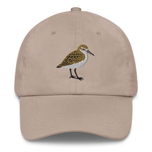 Puede incluir: Gorra de béisbol beige con un diseño de pájaro bordado. El pájaro tiene plumas marrones y blancas, un pico negro y patas negras. La gorra tiene una visera curva y una correa ajustable en la parte trasera.