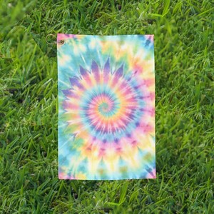 May include: A rectangular tie-dye towel with a spiral pattern in shades of blue, green, yellow, pink, and purple. The towel has a metal grommet in the upper left corner. The towel is lying on a bed of green grass.