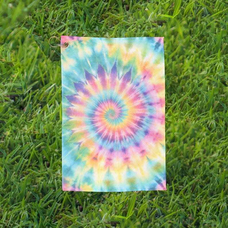 May include: A rectangular tie-dye towel with a spiral pattern in shades of blue, green, yellow, pink, and purple. The towel has a metal grommet in the upper left corner. The towel is lying on a bed of green grass.