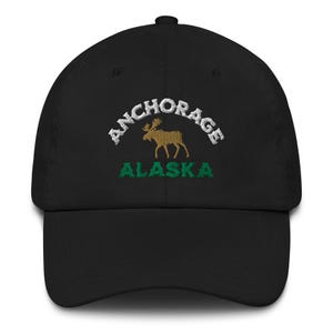 Könnte beinhalten: Schwarze Baseballkappe mit gesticktem Text und Grafik. Der Text lautet "ANCHORAGE" über einer goldenen Elchgrafik und "ALASKA" in Grün darunter. Eine klassische, verstellbare Kappe.