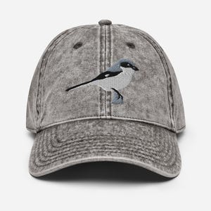 Puede incluir: Gorra de béisbol gris de estilo vintage con un diseño de pájaro bordado. El pájaro está representado en negro, blanco y gris, con un contorno detallado. La gorra tiene una visera curva y una correa ajustable.