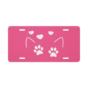 May include: A bright pink license plate featuring a white cat face design. The design includes two paw prints, two hearts, and cat ears. The plate has four mounting holes.