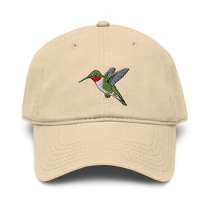 Puede incluir: Una gorra de béisbol beige con un bordado de un colibrí en vuelo. El ave presenta alas verdes, pecho blanco y una mancha roja brillante en la garganta, centrada en el panel frontal de la gorra estructurada de seis paneles.