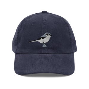 May include: Navy blue corduroy baseball cap featuring an embroidered bird design. The bird is depicted in shades of gray and white, with a black beak and eye. The cap has a curved brim and an adjustable strap at the back.