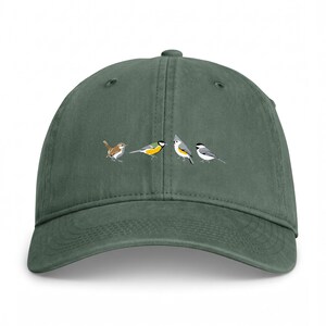 Peut inclure: Une casquette de baseball vert olive foncé ornée d'un imprimé graphique représentant quatre petits oiseaux alignés. Les oiseaux sont illustrés de profil avec un plumage détaillé dans des tons marron, jaune, blanc, gris et noir.