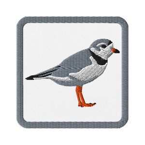 May include: Embroidered patch featuring a Piping Plover bird. The bird has gray wings and back, a white chest, and orange legs. It stands on a white background within a gray square border. The bird's beak is black and orange.