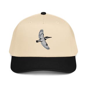 Puede incluir: Gorra de béisbol con corona color crema y visera negra. La gorra presenta un diseño bordado de un pájaro en vuelo, con detalles grises y negros. La gorra tiene un botón negro en la parte superior.