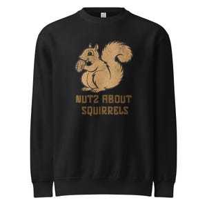 May include: Black sweatshirt with a tan squirrel graphic holding a nut. The text "NUTZ ABOUT SQUIRRELS" is printed below the squirrel in tan. The sweatshirt has a crew neck and long sleeves, suitable for cooler weather.