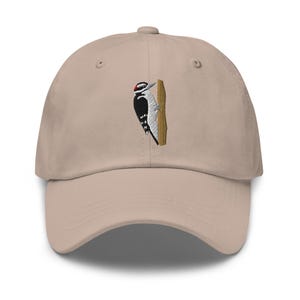 Könnte beinhalten: Eine beige Baseballkappe mit einer Stickerei eines schwarz-weiß-roten Spechtes, der auf einem braunen Baumstamm sitzt. Die Kappe hat einen gebogenen Schirm, eine strukturierte Krone mit Ösen und einen Knopf in der Mitte der Stoffkuppel.