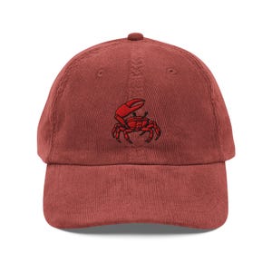 May include: A brick-red corduroy baseball cap featuring a red embroidered crab design on the front. The cap has a curved brim and an adjustable strap at the back. The crab is detailed with black outlines.