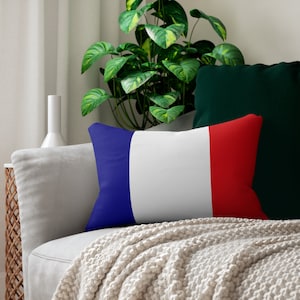 Könnte beinhalten: Ein weißes Sofa mit einer Wolldecke und zwei Kissen. Ein Kissen ist grün und das andere ist ein blau-weiß-rotes gestreiftes Kissen. Das gestreifte Kissen hat die Farben der französischen Flagge.